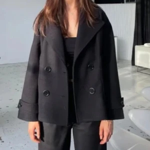 Svart kappa med knappar - Snygg svart kappa med dubbelknäppning och bred krage. Jackan har långa ärmar och en stilren design som passar perfekt för höst och vår. Perfekt för att ge en elegant touch till din outfit.