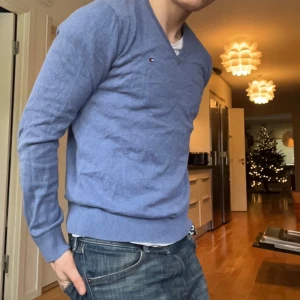 Blå tröja från Tommy Hilfiger - Säljer en stilren blå tröja från Tommy Hilfiger i bomull. Tröjan har en v-ringad design och långa ärmar, perfekt för en avslappnad look. Den lilla logotypen på bröstet ger en diskret touch av stil.