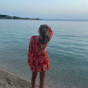 Röd och vit blommig klänning - Säljer en vacker röd och vit blommig klänning med volanger och öppen rygg. Klänningen har långa ärmar och en kort längd, perfekt för sommarens äventyr. Den har en omlott-design som ger en elegant touch.