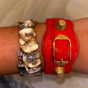 Rött armband från Balenciaga - Snyggt rött armband i skinn från Balenciaga med guldfärgade detaljer. Armbandet har en bred design med dekorativa nitar och ett spänne framtill. 