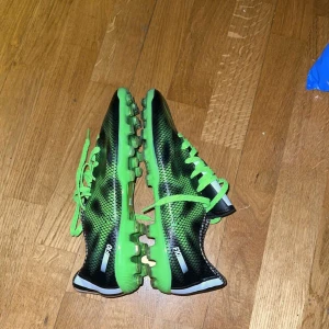 Gröna fotbollsskor från Adidas - Snygga gröna och svarta fotbollsskor från Adidas med ett häftigt mönster. Skorna har snörning och är perfekta för fotbollsplanen. De har en modern design med en kombination av gröna och svarta nyanser.