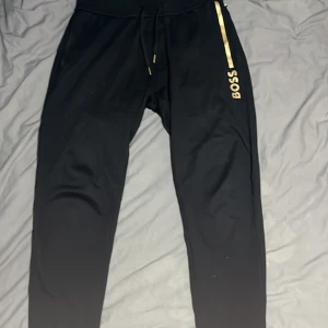 Svarta Hugo boss byxor  - Snygga svarta mjukisbyxor från Boss med gulddetaljer. Byxorna har en bekväm passform med dragsko i midjan och en stilren design med logotyp längs benet. Perfekta för en avslappnad look.