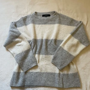 Grå och vit randig tröja från Vero Moda - Mysig stickad tröja från Vero Moda i grått och vitt med breda ränder. Tröjan har en normal passform och långa ärmar, perfekt för kyligare dagar.