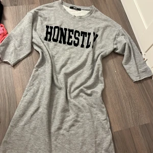 Grå klänning med text från Bik Bok - Snygg grå klänning från Bik Bok med texten 'HONESTLY' i svart på bröstet. Klänningen har trekvartsärmar och en avslappnad passform, perfekt för en casual look. Kortare ärmar