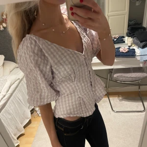 Rutig blus med volangdetaljer - Säljer en söt rutig blus i ljuslila och vit med volangdetaljer. Blusen har korta ärmar och knappar framtill. Perfekt för en somrig look!