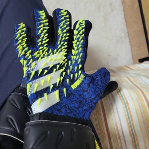 Blå och svarta fotbollshandskar - Säljer ett par coola fotbollshandskar i blått och svart med gula detaljer. Handskarna har en unik design med greppvänlig yta och är perfekta för målvakter. Märket är synligt på handskarna.