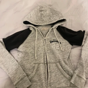 Grå och svart hoodie från Hollister - Säljer en snygg grå hoodie från Hollister med svarta ärmar och dragkedja. Den har en justerbar huva och fickor framtill. Perfekt för en avslappnad stil.