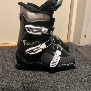 Svarta pjäxor från Salomon - Säljer ett par svarta pjäxor från Salomon med vita spännen och vadderad insida. Perfekta för vintersport och skidåkning. 