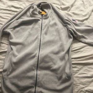 Grå tröja med dragkedja från Parajumpers - Säljer en stilren grå tröja från Parajumpers med dragkedja och ribbade detaljer. Tröjan har en broderad logga på ärmen och är perfekt för kyligare dagar. Den är tillverkad i ett mjukt material som ger en bekväm passform.