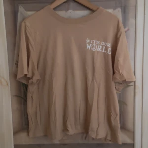 Beige t-shirt från H&M  - Säljer en beige t-shirt från H&M Divided med texten 'It's Our World' tryckt i vitt på både fram- och baksidan. T-shirten har en normal passform och korta ärmar, perfekt för en avslappnad stil.