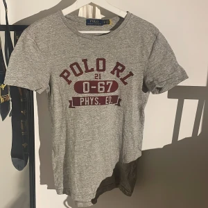Grå t-shirt från Polo Ralph Lauren - Snygg grå t-shirt från Polo Ralph Lauren med röd text som är helt oanvänd'POLO RL 0-67 PHYS. ED.' på framsidan. Perfekt för en avslappnad stil. Kortärmad och tillverkad i mjukt material. .nypris 1200kr 