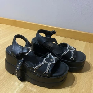 Svarta platåsandaler med hjärtdekoration - Snygga svarta platåsandaler med silverfärgade kedjor och hjärtformade detaljer. Sandalerna har en justerbar rem med spänne och en hög platåsula för extra höjd och stil.