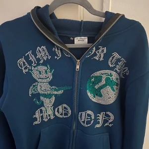 Blå hoodie med strassdetaljer från Aim for the Moon - Snygg blå hoodie från Aim for the Moon med glittrande strassdetaljer och dragkedja. Perfekt för att sticka ut med stil. Den har en cool design med text och motiv framtill. Grymt skick 9/10 pris 599 för att den ej säljs längre 