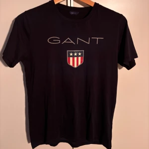 Mörkblå t-shirt från GANT - Snygg mörkblå t-shirt från GANT med tryck av logotyp på framsidan. Perfekt för en stilren och avslappnad look. Knappt använd. Storlek 158/164 (13-14) men passar perfekt för vuxen XS.