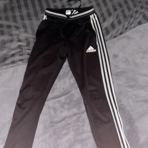 Svarta träningsbyxor från Adidas - Säljer ett par svarta träningsbyxor från Adidas med de klassiska tre vita ränderna längs sidorna. Byxorna har en elastisk midja med snörning och Adidas-loggan på låret. Perfekta för träning eller en avslappnad stil. Inga defekter men dem är väl använda