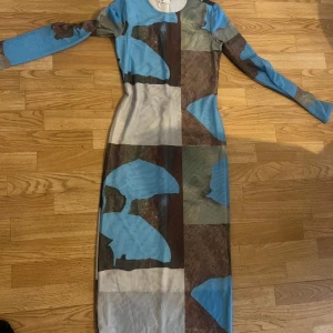 Mönstrad långklänning från Monki - Säljer en cool långklänning från Monki med ett unikt mönster i blått, brunt och grönt. Klänningen har långa ärmar och en figurnära passform. Perfekt för dig som vill sticka ut med en färgglad och modern stil. Har endast använts en gång, mycket bra skick.