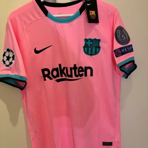 Rosa FC Barcelona fotbollströja - Säljer en rosa FC Barcelona fotbollströja från Nike med korta ärmar. Tröjan har klubbens emblem och sponsorlogga på framsidan samt Messi och nummer 10 på baksidan. Detaljer i svart och turkos vid ärmslut och krage. Perfekt för fans av klubben!
