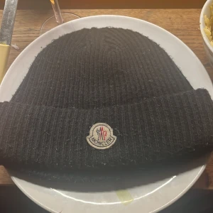 Svart ribbad mössa från Moncler - Snygg svart ribbad mössa från Moncler med broderad logotyp framtill. Perfekt för att hålla värmen under kyliga dagar. Klassisk design som passar till det mesta.