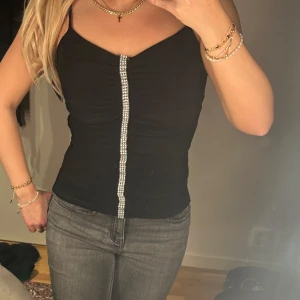 Svart linne med strassdetalj - Snygg svart linne med smala axelband och en glittrig strassdetalj framtill. Perfekt för en kväll ute eller en festlig tillställning. Enkel och stilren design som passar till det mesta.