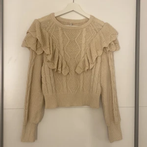 Beige stickad tröja med volangdetaljer - Säljer en beige stickad tröja med volangdetaljer på axlarna. Tröjan har ett flätmönster och långa ärmar. Bra skick andvänt 1-2 gånger.