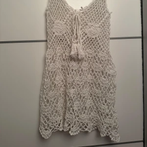 Vit virkad cover up klänning - Charmig vit virkad klänning med knytning framtill. Perfekt för sommarens varma dagar med sin luftiga design och vackra detaljer. Klänningen har en kort längd och breda axelband.