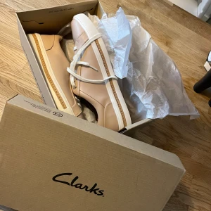 Beige sneakers från Clarks - Snygga beige-ros sneakers från Clarks med vit sula och snörning. Perfekta för en stilren look. Skorna har en enkel och modern design som passar till många olika outfits.Helt nya med kartong 
