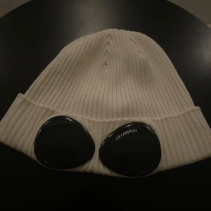 Vit Cp company mössa - Snygg vit ribbad mössa från C.P. Company med unika glasögondetaljer framtill. Perfekt för att hålla stilen under kyliga dagar. Tillverkad i 100% bomull för en bekväm passform. Jag säljer pga att den är lite stor för mig. 