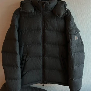 Grå dunjacka från Moncler - Säljer en snygg grå dunjacka från Moncler med dragkedja och hög krage. Jackan har långa ärmar och en broderad logotyp på ärmen. Perfekt för kyliga dagar.