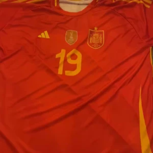 Röd fotbollströja från Adidas - Säljer en röd fotbollströja från Adidas med gula detaljer och nummer 19. Tröjan har korta ärmar och ett emblem på bröstet. Perfekt för fotbollsfans!