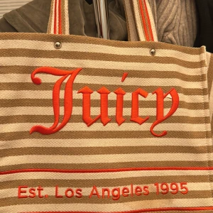 Juicy Couture shoppingväska/strandväska - Snygg randig shoppingväska eller strandväska från Juicy Couture i beige och rött. Väskan har texten 'Juicy' och 'Est. Los Angeles 1995' broderad på framsidan. Insidan är mönstrad med Juicy Couture-logotypen och har flera praktiska fack med dragkedja. Nypris ca 1000kr men säljer för 400kr. Helt helt oanvänd!