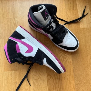Air Jordans 1 Mid Magenta  - Nike Air Jordans 1 Mid Magenta i en färgkombination av svart, vitt och rosa. Skorna har en klassisk hög modell med snörning och den ikoniska Air Jordan-loggan på sidan. Använda två gånger så som nya i skicket. Köptes för 5000kr.