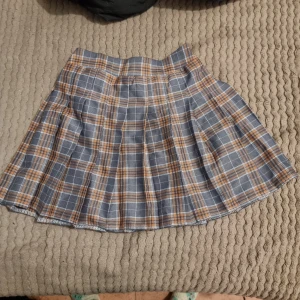 Rutig kjol i grått och orange - Snygg rutig kjol i grått och orange med en klassisk design. Kjolen har en kort längd och är perfekt för en trendig look. Den har en bekväm passform och är lätt att matcha med olika toppar. Storleken är uppskattad, den är 140 i barnstorlek men passar xxs och xs.