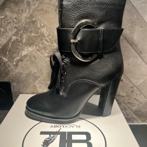 Svarta boots med snörning och bälte från Black Glory - Snygga svarta boots med hög klack och snörning framtill. Skorna har en bred rem med ett stort, runt spänne som ger en cool detalj. Perfekta för att ge din outfit en edgy touch. Sparsamt använda. Inköpta i Danmark för 1399:-DKK.