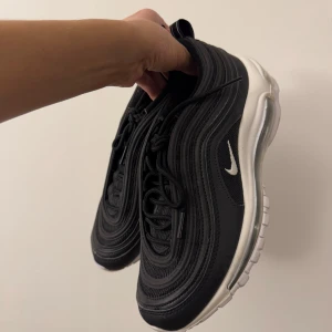 Svarta Nike Air Max sneakers - Snygga svarta Nike Air Max sneakers med vit sula och ikonisk synlig luftdämpning. Skorna har en elegant design med snörning och den klassiska Nike-loggan i vitt på sidan.