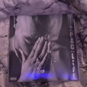 Best of 2Pac - Life Part 2 Vinyl - Säljer ett album av 2Pac med titeln 'Life Part 2'. Omslaget har en svartvit bild av artisten med händerna i bön. Baksidan visar en bild av honom utan tröja och låtlistan. Perfekt för hiphop-fans och samlare! Aldrig använd!