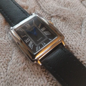 Svart klocka med läderarmband, helt ny  - Elegant klocka med svart urtavla och romerska siffror. Den har en rektangulär boett i silver och ett svart läderarmband. Perfekt för en stilren look.