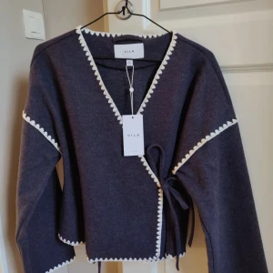 Mörkblå omlottkofta från Vila - Snygg mörkblå omlottkofta från Vila med vita spetsdetaljer längs kanterna. Koftan har långa ärmar och knytband i sidan för en justerbar passform. Perfekt för en stilren look.