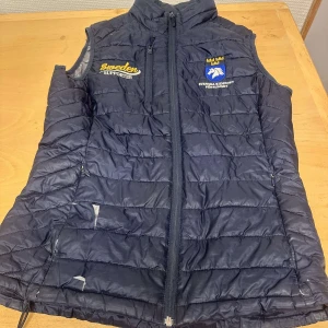 Mörkblå quiltad väst från svensk ridsport  - Säljer en mörkblå väst från svensk ridsport med dragkedja framtill. Västen har broderade detaljer med texten 'Sweden Supporter' och en logotyp på bröstet. Perfekt för sportiga tillfällen eller som en stilren accessoar. Ett litet hål