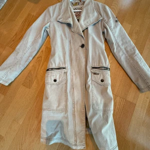 Beige trenchcoat från Khujo - Snygg beige trenchcoat från Khujo med långa ärmar och knappar framtill. Jackan har två stora fickor med knappar och dragkedjor samt ett bälte med texten 'Genuine' för en cool look. Perfekt för vår och höst!