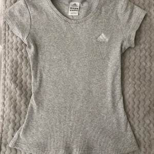Grå ribbad t-shirt från Adidas - Säljer en vintage-stilren grå ribbad t-shirt från Adidas med korta ärmar och rund hals. Perfekt för en sportig look eller avslappnad stil. Adidas-loggan är diskret placerad på bröstet. Tight fit och väldigt nice!🦦