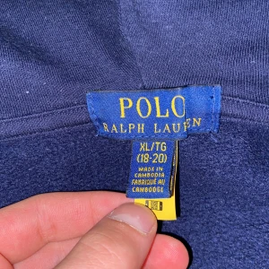 Mörkblå hoodie från Ralph Lauren storlek 170/88. - Säljer en snygg mörkblå hoodie från Ralph Lauren med dragkedja och huva. Den har en broderad röd logotyp på bröstet och är perfekt för en avslappnad stil. Passar bra till både jeans och joggers. Hör av dig vid intresse! 