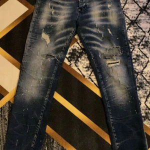 Blå dsq jeans - Säljer ett par blå jeans med slitningar och en cool, tvättad look. De har en klassisk femficksdesign och en bekväm passform. Perfekta för en avslappnad stil.