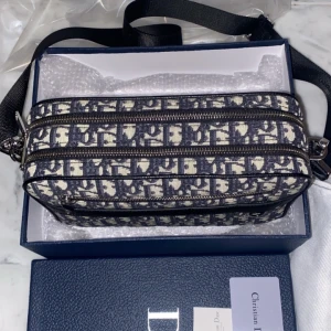 Mönstrad axelväska från Dior - Snygg axelväska från Dior med ett ikoniskt mönster i svart och beige. Väskan har flera dragkedjor och en justerbar axelrem. Perfekt för att bära dina nödvändigheter med stil. Oanvänd!