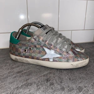Golden Goose - Säljer ett par glittriga sneakers från Golden Goose i väldigt bra skick 9/10 använda 1-4 gånger. Nypris 5400-7000kr vårt pris 1099kr. Passar 39/40 rekommenderar att man lägger i en extra sula om man har 39. Dem slitningar som syns är en del av designen då det ska se ut som att man skateat dem. Skorna har en färgglad glittrig yta med gröna detaljer på hälen. Perfekta för att ge din outfit en extra touch! Vid behov av fler bilder eller allmänna frågor tveka inte på att skriva till oss 😁💚