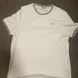 Vit t-shirt från Slazenger - Säljer en klassisk vit t-shirt från Slazenger med korta ärmar och rund halsringning. T-shirten har en diskret logga på bröstet och kontrasterande ränder vid hals och ärmslut. Perfekt för en avslappnad stil. Avhämtning i Göteborg eller Mölndal. Ingen frakt!