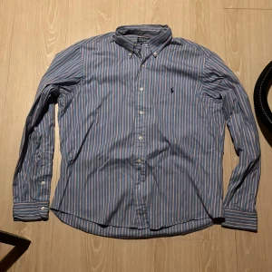 Randig skjorta från Polo Ralph Lauren - Säljer en stilren randig skjorta från Polo Ralph Lauren i blått, vitt och rött. Skjortan har långa ärmar och knappar framtill samt en broderad logga på bröstet. Perfekt för en klassisk look!