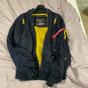 Svart bomberjacka från Alpha Industries - Säljer en snygg svart bomberjacka från Alpha Industries med gul insida. Jackan har långa ärmar och dragkedjor i gult. Den ikoniska 'Remove Before Flight'-taggen finns på ärmen. Väldigt skön jacka i denna årstid men passar även bra i andra väder. Den är väldigt välbehandlad så den är i bra skick och har inga hål eller andra märken. 