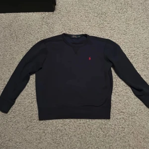 Mörkblå tröja från Polo Ralph Lauren - Säljer en mörkblå tröja från Polo Ralph Lauren med det klassiska röda logotypbroderiet på bröstet. Tröjan har långa ärmar och en rund halsringning. 