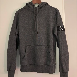 Grå hoodie från Calvin Klein - Säljer en stilren grå hoodie från Calvin Klein med en klassisk logga på ärmen. Tröjan har en känguruficka och justerbar huva med dragsko. Perfekt för en avslappnad look.