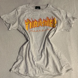Vit Thrasher t-shirt med flamtryck - Säljer en vit t-shirt från Thrasher med det ikoniska flamtrycket i rött och gult. Perfekt för skate-entusiaster eller streetwear-fans. T-shirten har en klassisk passform och är kortärmad.